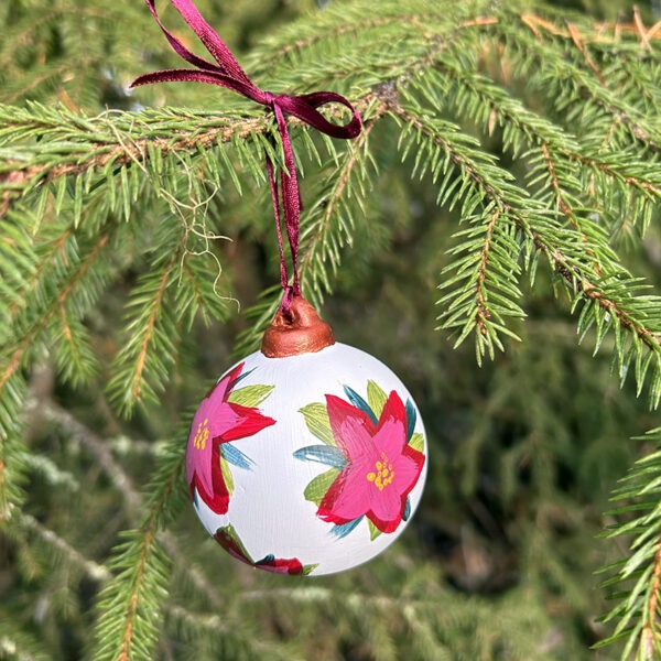 Bilde av håndmalt julekule hengende i et juletre. Julekulen er hvit med blomsten julestjerne